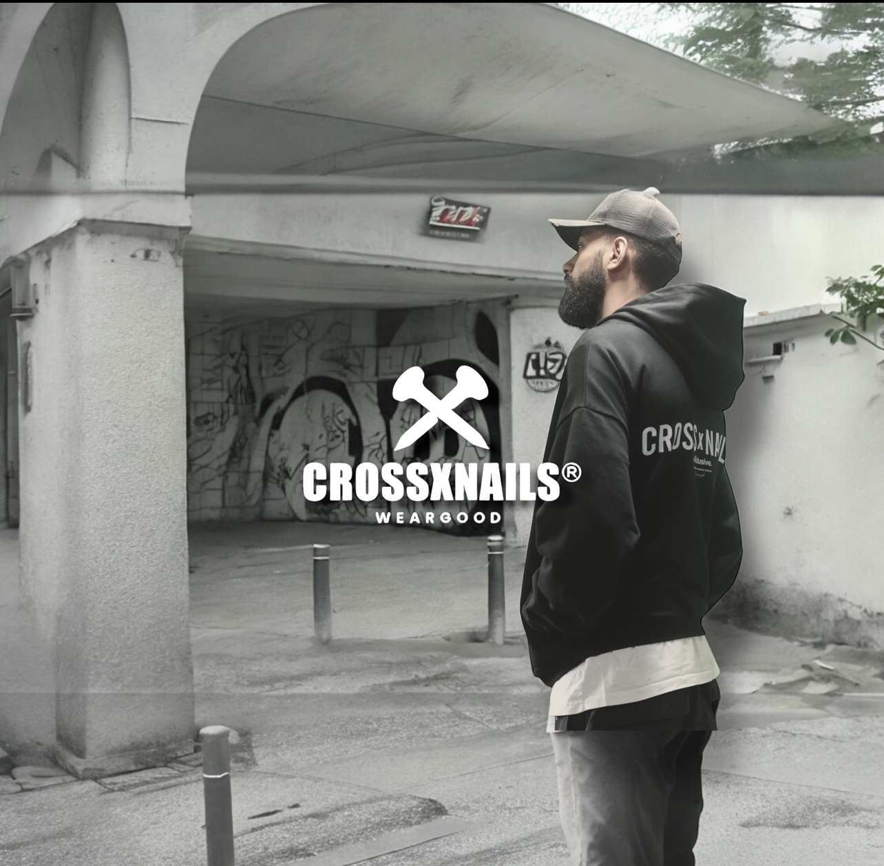 A male model wearing a black oversized Crossxnails® boxy fit hoodie with a minimalist white Crossxnails® exclusive icon on the back. He stands on an urban street, facing sideways, showcasing the hoodie’s back design. Part of the “On a Mission” collection.